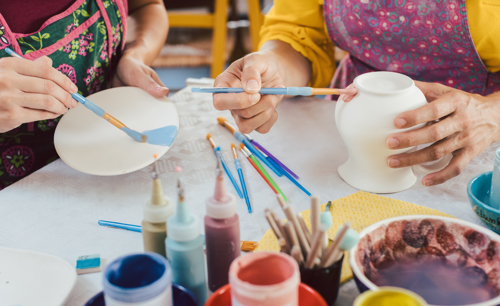 Picture of two people's hands painting a pottery jug and plate in different colours